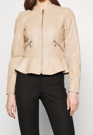 Veste beige en simili cuir zippée, avec bas à basque, poches zippées en biais et manches longues, portée avec un pantalon noir.