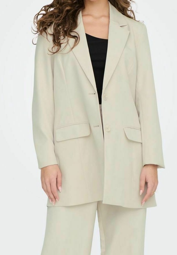 Femme portant un blazer beige et un pantalon assorti sur un haut noir, debout devant un fond blanc uni.