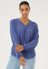 Langsleeve, V-hals blouse in koningsblauw, gemaakt van textuurstof. Heeft gathered mouwen en een ontspannen pasvorm, gedragen met lichte denim.