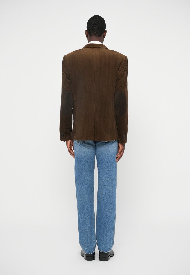 Brun kordfløyelsblazer med axelvaddar och armbågsplåster, parat med ljusblå raka jeans och svarta skor.