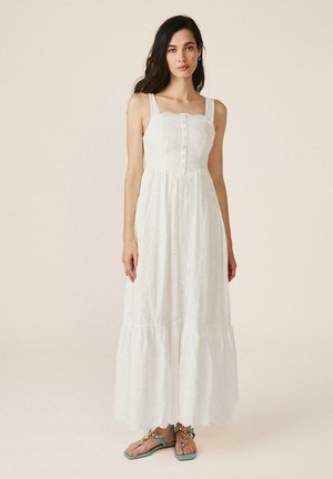 Mujer con cabello largo y oscuro, vestida con un vestido blanco sin mangas de ojales y sandalias verdes adornadas, de pie frente a un fondo liso.