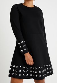 Musta mekko pitkillä hihoilla, jossa on ympyränmuotoisia hopeisia niittejä helman ja hihojen resorien ympärillä, valmistettu teksturoidusta kankaasta.