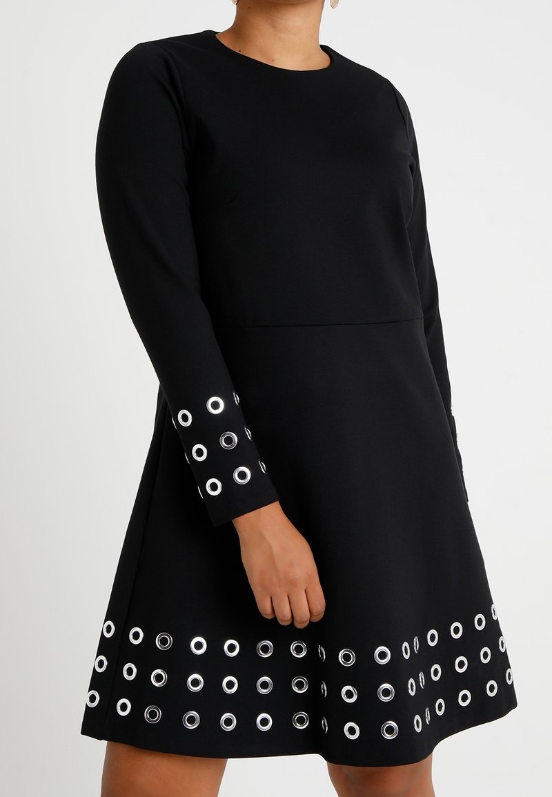 Musta mekko pitkillä hihoilla, jossa on ympyränmuotoisia hopeisia niittejä helman ja hihojen resorien ympärillä, valmistettu teksturoidusta kankaasta.