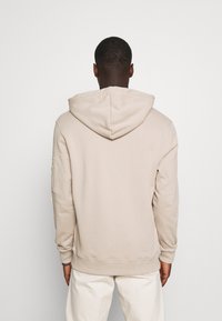 Bēša kapuce ar elastīgu kapuci, ribinātām piedurknēm un kabatu. Mīksts audums ar brīvu piegriezumu un bezšuvju aizmugures dizains.