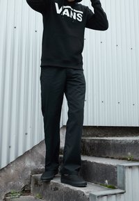 Sudadera negra con el logo blanco de Vans, combinada con pantalones negros y zapatillas sin cordones. De pie sobre escaleras de piedra gris con un fondo de chapa.
