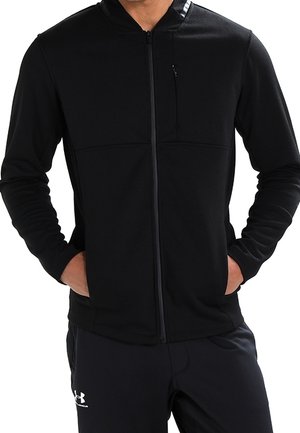 Mężczyzna w czarnej bluzie z kapturem zapinanej na zamek, z rękami w kieszeniach i czarnych spodniach Under Armour, wykadrowany poniżej nosa.