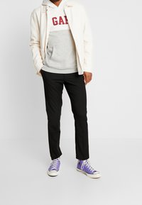Conjunto de hombre que incluye una chaqueta crema, sudadera gris y blanca con texto rojo, pantalones negros y zapatillas moradas con suelas blancas.