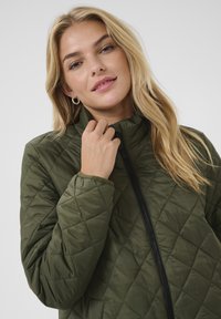 Veste matelassée vert olive avec un col haut, une fermeture éclair noire et un tissu légèrement texturé. Présente un motif en losanges et des manches ajustées.
