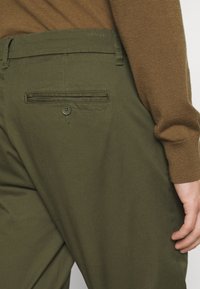 Pantaloni verde oliva con una superficie liscia, dotati di una tasca sul retro con chiusura a bottone e un sottile design piegato in vita.