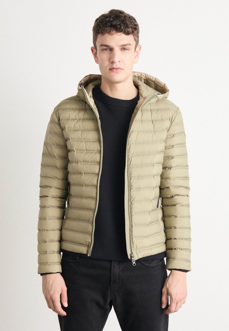 Svetlo zelena napihnjena jopica s kapuco, z vodoravnimi prešitimi deli, sprednjim zadrgo in stranskimi žepi, oblečena čez temno pulover.