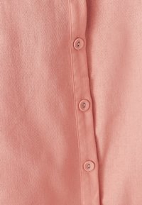 Breskva košulja s dugim rukavima od teksturiranog materijala s pet usklađenih dugmeta duž plaveta i glatkom završnom obradom.