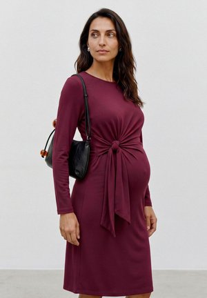 Gravid kvinde i en langærmet vinrød kjole med knyte-detalje foran, bærende en sort skuldertaske, stående mod en ensfarvet baggrund.