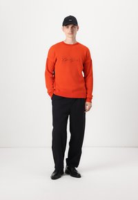 Pullover arancione a costine con logo ricamato, abbinato a pantaloni neri e scarpe nere. Il modello indossa un berretto nero. Sfondo minimale.