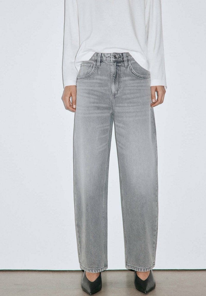 Personne portant un jean large gris clair, une chemise blanche à manches longues et des chaussures noires à bout pointu, debout devant un fond blanc.