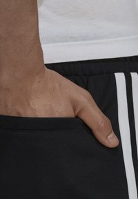 Černé sportovní šortky s bílými pruhy, texturovaná látka, na boku s rukou položenou v kapsě. Nahoře viditelné bílé tričko.