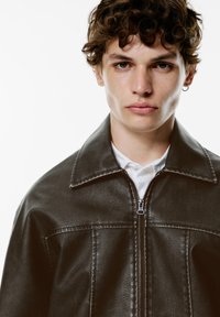 Veste en cuir synthétique marron foncé avec fermeture éclair à l'avant, col pointu et texture doublée ; portée sur une chemise blanche. Portrait en gros plan d'un jeune homme.