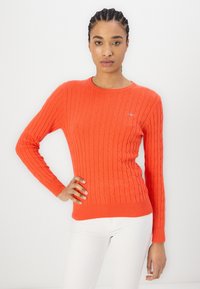 Suéter naranja de punto trenzado con cuello redondo y mangas largas. Detalle de puños y dobladillo acanalados. Llevado con pantalones blancos.