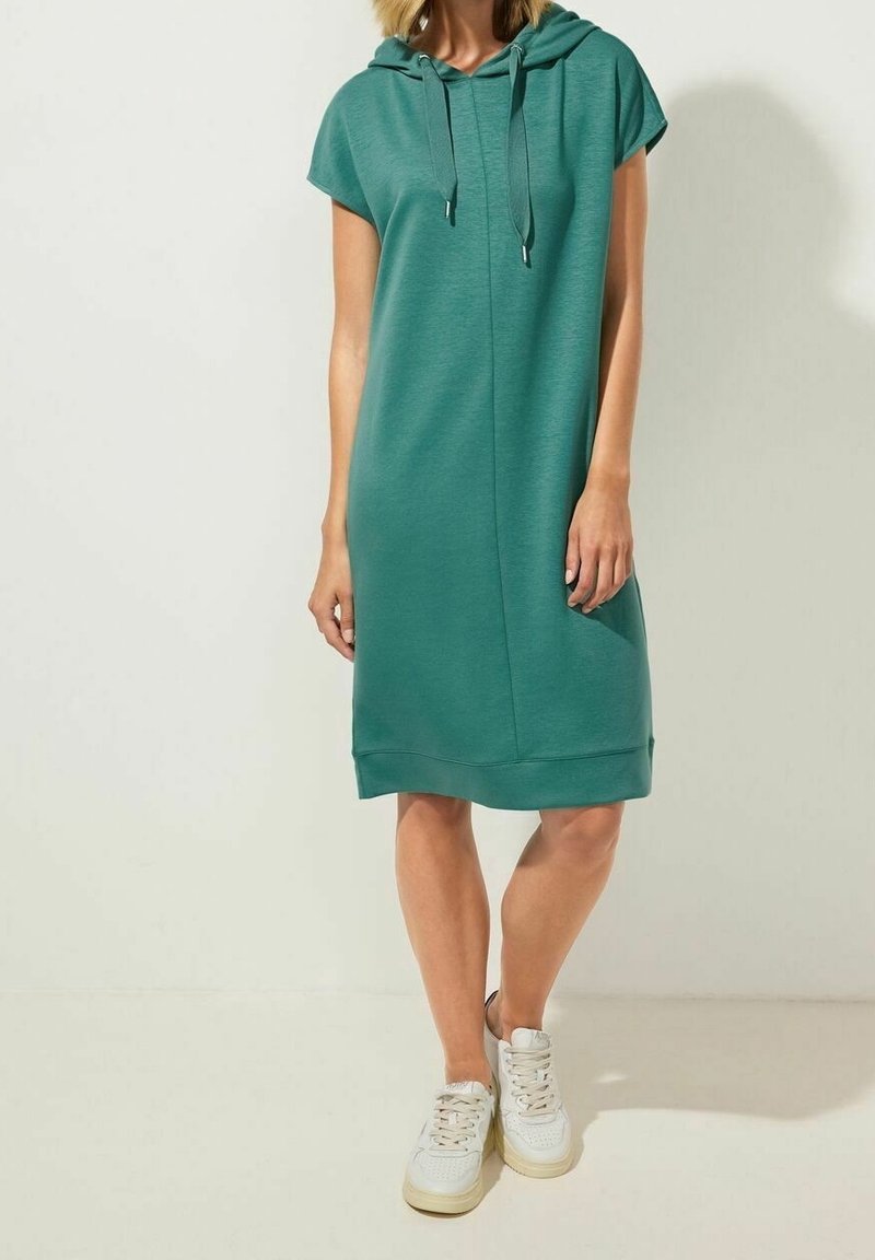 Groene hoodie-jurk met korte mouwen, gestructureerde stof, losse pasvorm en zijzakken, gecombineerd met witte sneakers.