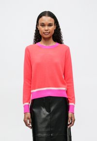 Mujer con cabello rizado que lleva un suéter coral rosa con ribetes en rosa brillante y beige, combinado con una falda de piel negra, de pie contra un fondo blanco.