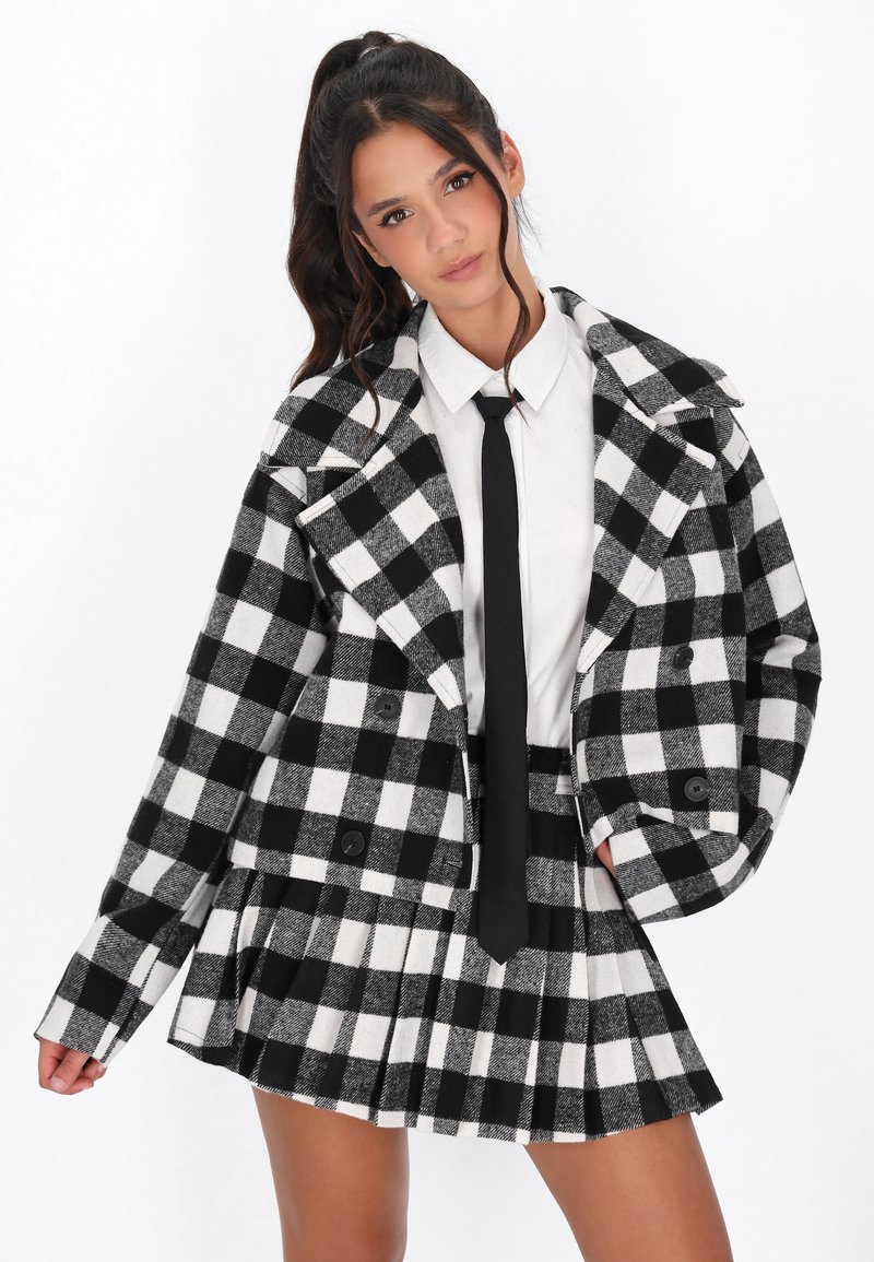 Cappotto a scacchi bianco e nero con ampi rever e bottoni doppi, abbinato a una gonna a pieghe con scacchi e una camicia bianca.