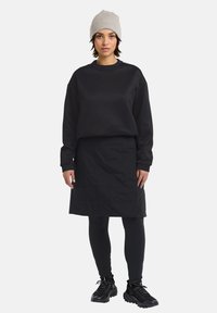 Felpa nera con collo rotondo, abbinata a una gonna nera trapuntata. Leggings scuri e sneakers nere texturizzate. Beanie lavorato a maglia grigio chiaro in testa.