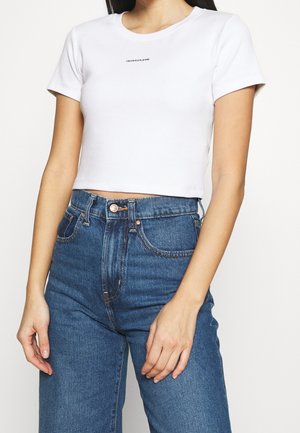Kvinde iført en hvid cropped Calvin Klein Jeans t-shirt og højtaljede blå denimjeans, stående foran en ensfarvet baggrund.