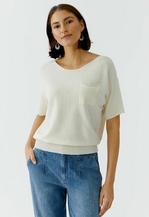 Frau trägt cremefarbenes Kurzarm-Strickoberteil mit Brusttasche und blaue High-Waist-Jeans, steht mit einer Hand in der Tasche vor weißem Hintergrund.