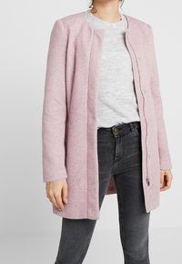 Lichtroze wolmixjas met een recht ontwerp, lange mouwen en een drukknopen sluiting. Gemaakt over een grijze trui en donkere jeans.