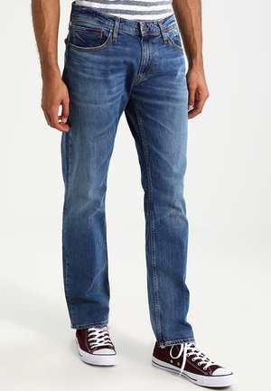 Homme portant un jean bleu, une chemise rayée et des baskets rouge foncé, debout sur un fond blanc.