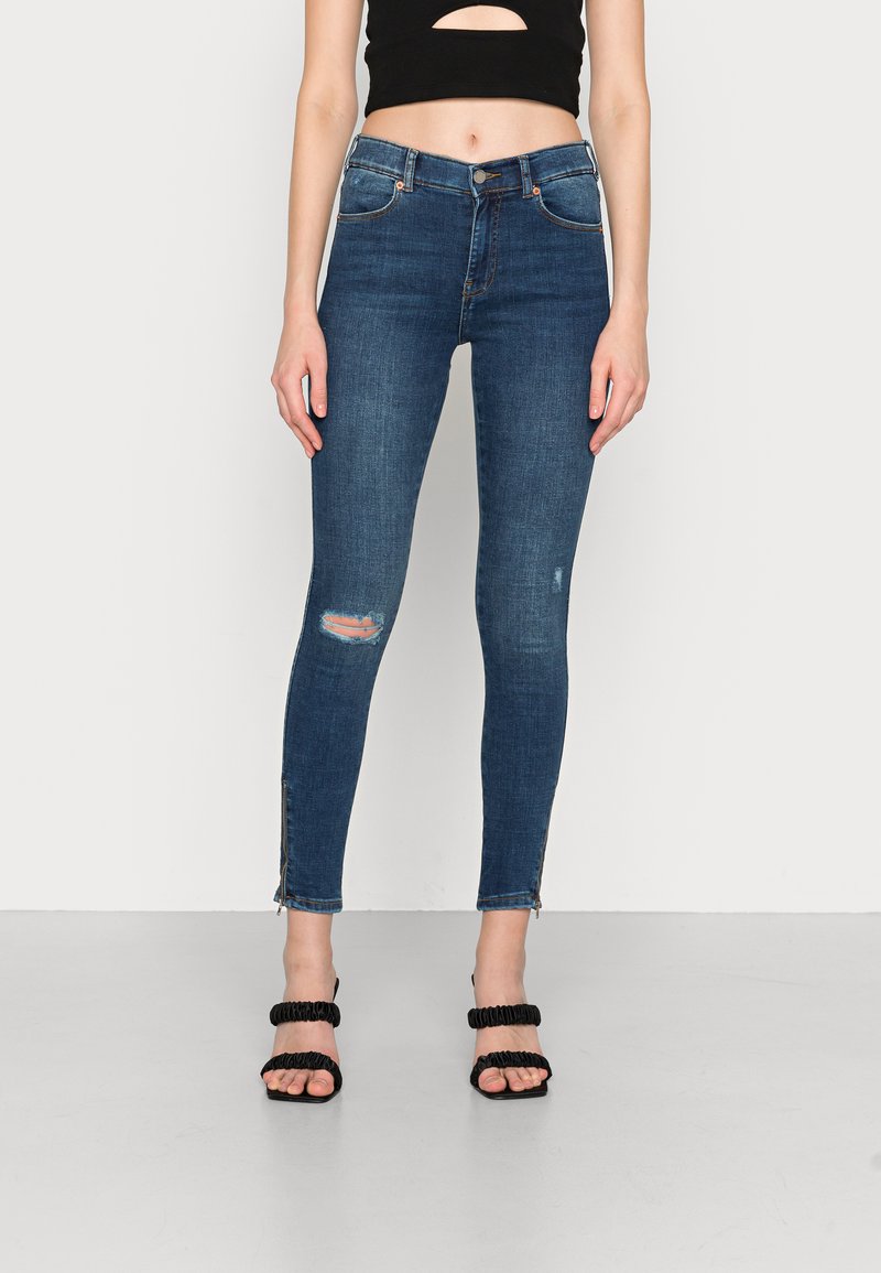 Jeans skinny em denim azul escuro, com cortes rasgados nos joelhos e zíperes nos tornozelos. Combinados com saltos altos pretos com tiras e um top cropped.