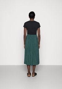 Top nero a maniche corte abbinato a una lunga gonna verde plissettata. Sandali neri piatti con suola testurizzata. Sfondo neutro.