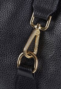 Dettaglio di una borsa in pelle nera con gancio a moschettone color oro e anello circolare. Superficie testurizzata con una delicata trama.