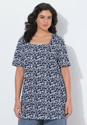 Frau mit dunklem lockigem Haar, die eine weiße Tunika mit marineblauem Blumenmuster und kurzen Ärmeln trägt, vor einem schlichten hellen Hintergrund stehend.