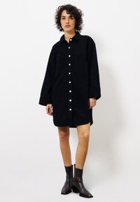 Schwarzes Hemdkleid mit Kragen, zwei Brusttaschen und langen Ärmeln. Getragen mit braunen Knöchelstiefeln. Glatte Stofftextur.