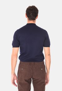 Uomo in piedi di spalle, indossa una camicia navy a maniche corte e pantaloni in corduroy marrone con tasche posteriori a bottone.