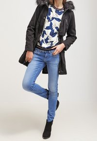 Osoba ubrana w czarną kurtkę z kapturem i futrzanym wykończeniem, białą bluzę z niebieskim kwiatowym wzorem, jasnoniebieskie dżinsy i czarne botki za kostkę, stojąca z jedną nogą skrzyżowaną.