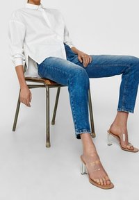 Klart klackade sandaler med en transparent rem, beige sula, bärs med blå denimjeans och en vit skjorta med knappar, sittande på en stol.