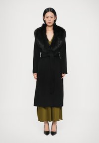 Garš melns mētelis ar kažokādas apkakli, jostots viduklī, uz olīvu zaļa kleita. Modelis valkā melnas kurpes ar smailiem purngaliem.