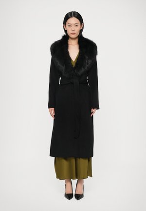 Lange zwarte jas met een bontkraag, in de taille gestrikt, over een olijfgroene jurk. Het model draagt zwarte pumps met een puntige neus.
