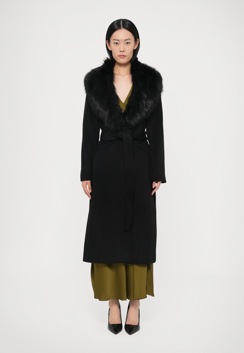 Garš melns mētelis ar kažokādas apkakli, jostots viduklī, uz olīvu zaļa kleita. Modelis valkā melnas kurpes ar smailiem purngaliem.