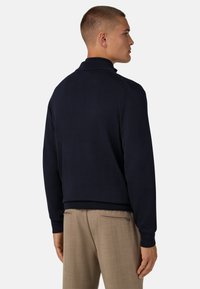 Mėlynas megztinis su aukštu apykakle, raštuotomis rankovėmis ir prigludusia forma, derinamas su šviesiais smėlio spalvos kelnėmis su užtrauktukais kišenėse.