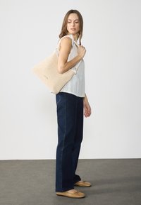 Beżowa pleciona torba z fakturowanym wykończeniem, o luźnym fasonie. Modelka nosi jasną bluzkę i ciemnoniebieskie dżinsy, z brązowymi płaskimi butami.