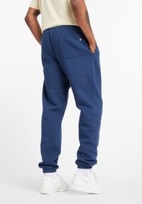 Mann trägt blaue Jogginghosen mit elastischen Bündchen und weiße Sneakers, steht mit den Händen in den Taschen vor einem weißen Hintergrund.