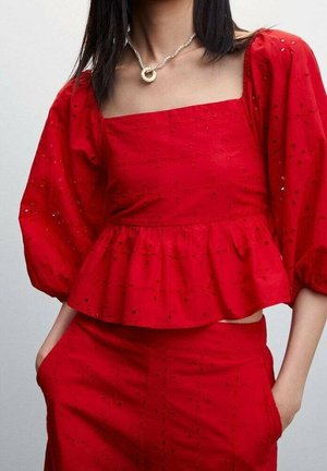 Mujer con blusa peplum de manga abullonada roja y pantalones a juego, con las manos en los bolsillos, complementada con un collar de perlas con colgante circular.