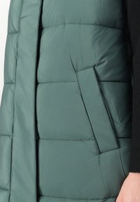 Grønn polstret vest med horisontalt quiltet mønster, sidelomme og glatt tekstur. Plagget har høy krage og er lagdelt over en svart topp.