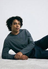 Pelēka garām piedurknēm tita krekls ar apaļu izgriezumu un Calvin Klein logo neuzkrītošā krāsā. Modelis demonstrē brīvu piegriezumu kopā ar jūraszilu svītrainām biksēm.