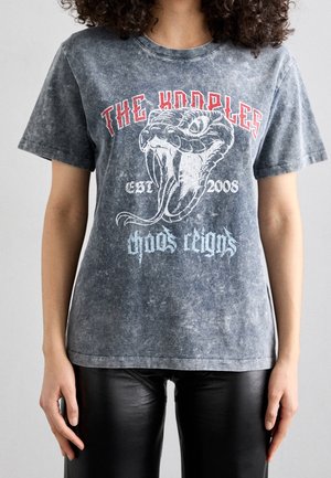 Person trägt ein graues ausgebleichtes T-Shirt mit einer großen Schlangengrafik und den Texten "The Kooples", "est. 2008" und "chaos reigns."