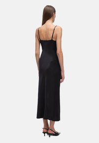 Kobieta z prostymi brązowymi włosami ma na sobie czarną bezrękawnikową sukienkę midi i czarne buty na niskim obcasie typu slingback, stojąc tyłem.