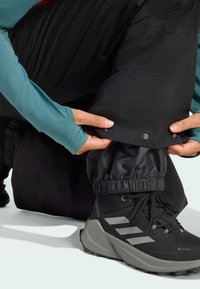 Čierne vodoodpudivé nohavice s elastickými manžetami, s detailom z kovových cvočkov, skombinované s čiernymi a šedými topánkami GORE-TEX s textúrovanými podrážkami.