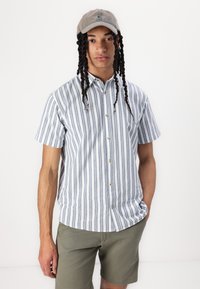 Chemise à manches courtes avec des rayures verticales bleues et blanches, boutons sur le devant, associée à un short vert olive et une casquette beige.